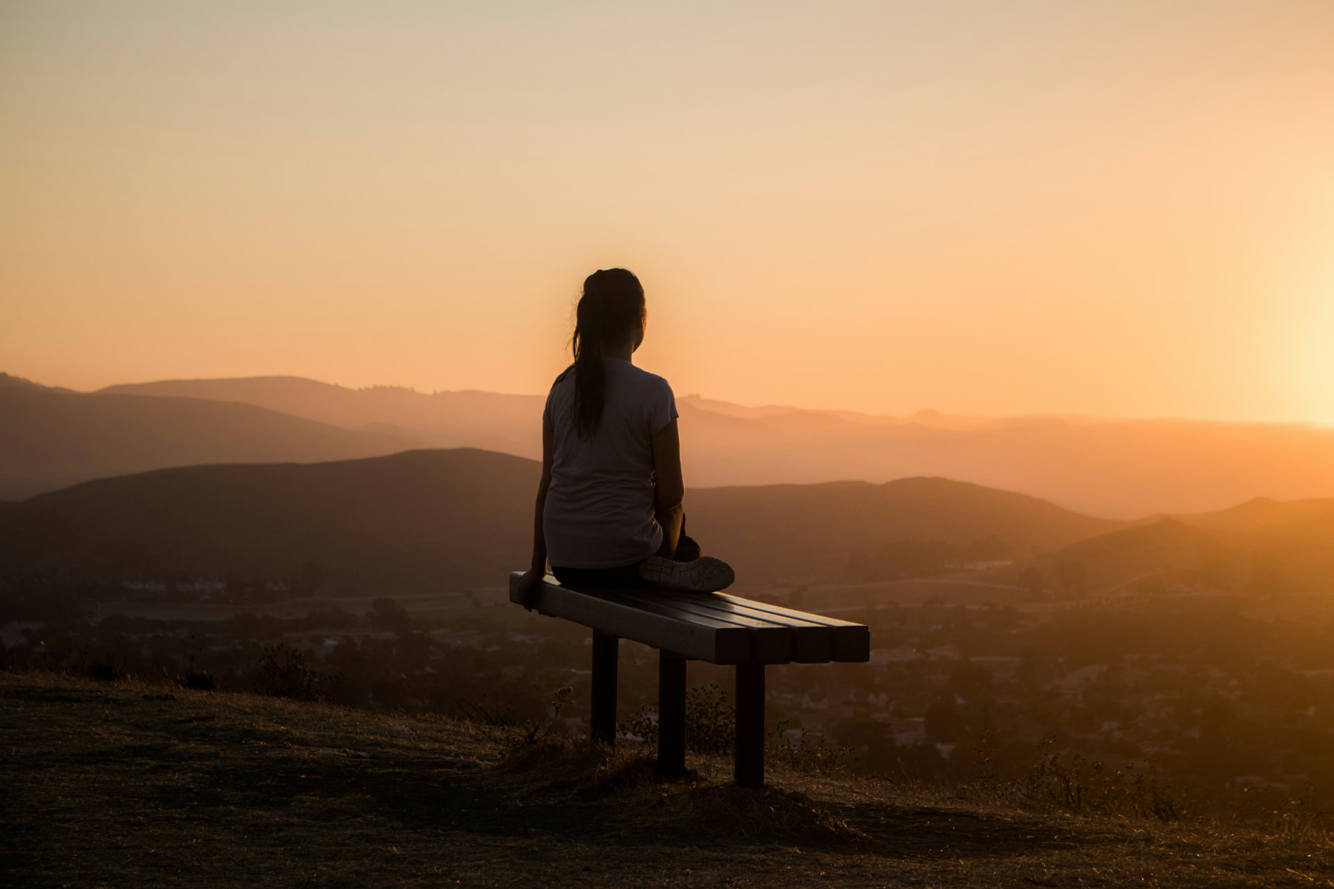 A person sitting on a hilltop bench at sunset, looking out over rolling California hills