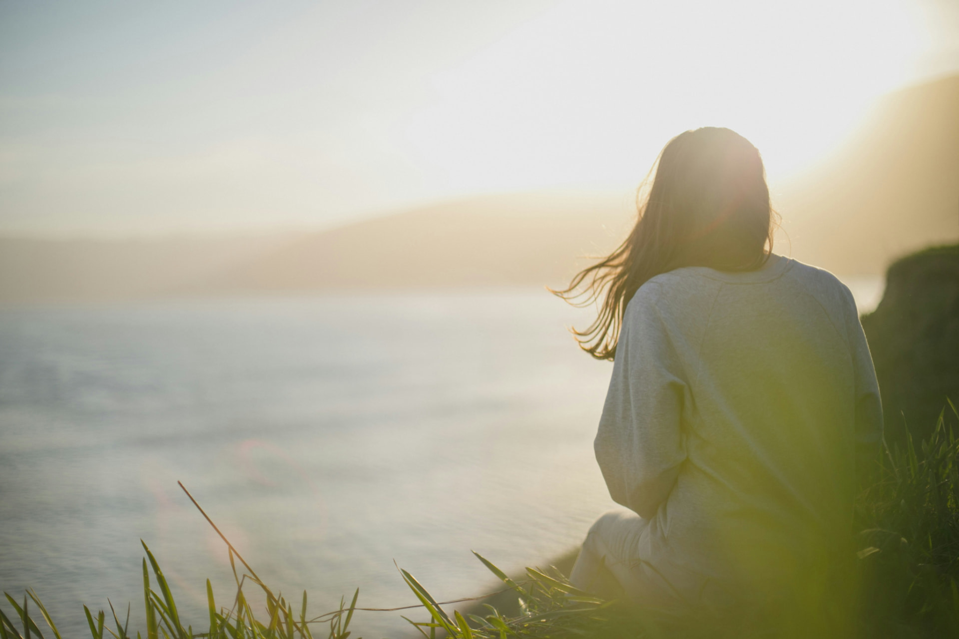 A woman sitting on a coastal bluff at golden hour, looking out over the water